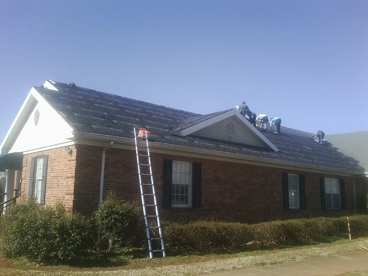 Crew tearing shingles off a brick ranch roof with a ladder set — Emergency Roof Repair work by our crew in Lacombe