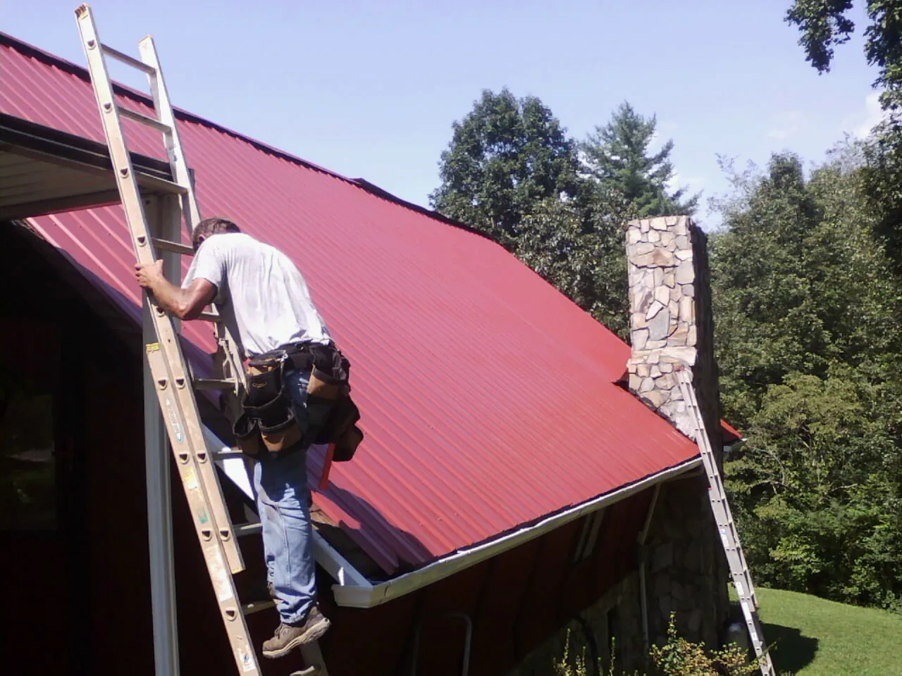 Roofer on a ladder beside a red metal roof and stone chimney — Soffit & Fascia Repair by Roofing Brothers in Lacombe